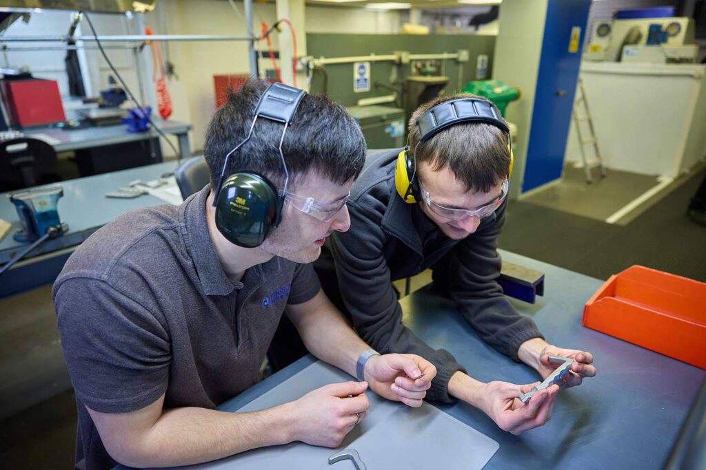 Kirkstall Precision Engineering 0171 - Two men lean over a table and look at a small metal part, man on the left wears black ear defenders, man on the right has yellow defenders, they are both looking down at the part, an orange tray sits to middle right of the image.