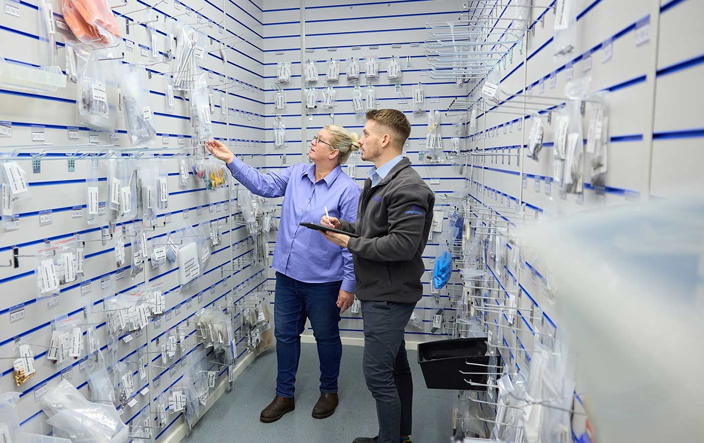 Nina and Matt in the Kirkstall Precision store room - A man and a woman in a white store room with many packaged products hanging on the walls. The woman has right hand outstretched to hold a packet. The man holds a stylus and clipboard.